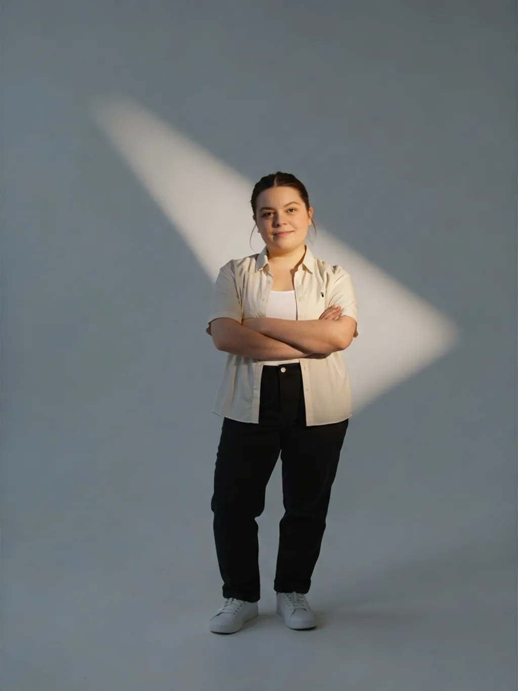 Cream short-sleeve shirt open over a white vest, black straight-leg jeans and white trainers, arms folded, on a grey backdrop